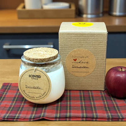 Scented candle in a glass jar with a cork lid on a red plaid cloth, next to a cardboard box and an apple.