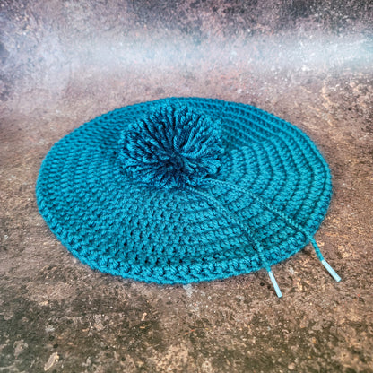 Teal crocheted beret with pom-pom, flat lay.