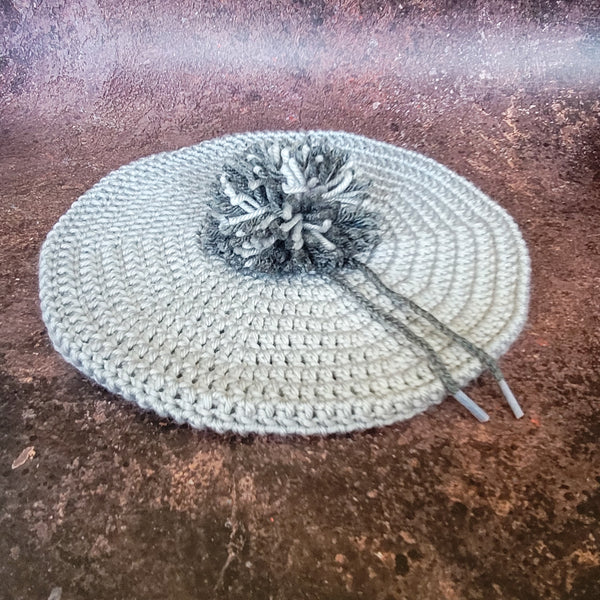 Two-toned grey crochet beret with detachable pompom, flat lay.