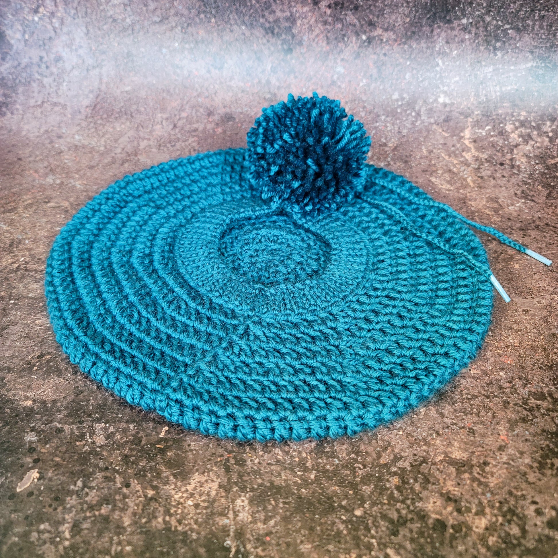 Teal crocheted beret with pom-pom on side, flat lay.