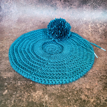 Teal crocheted beret with pom-pom on side, flat lay.