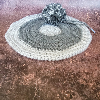 Flat lay of two-toned grey crochet beret with detachable pompom on side.