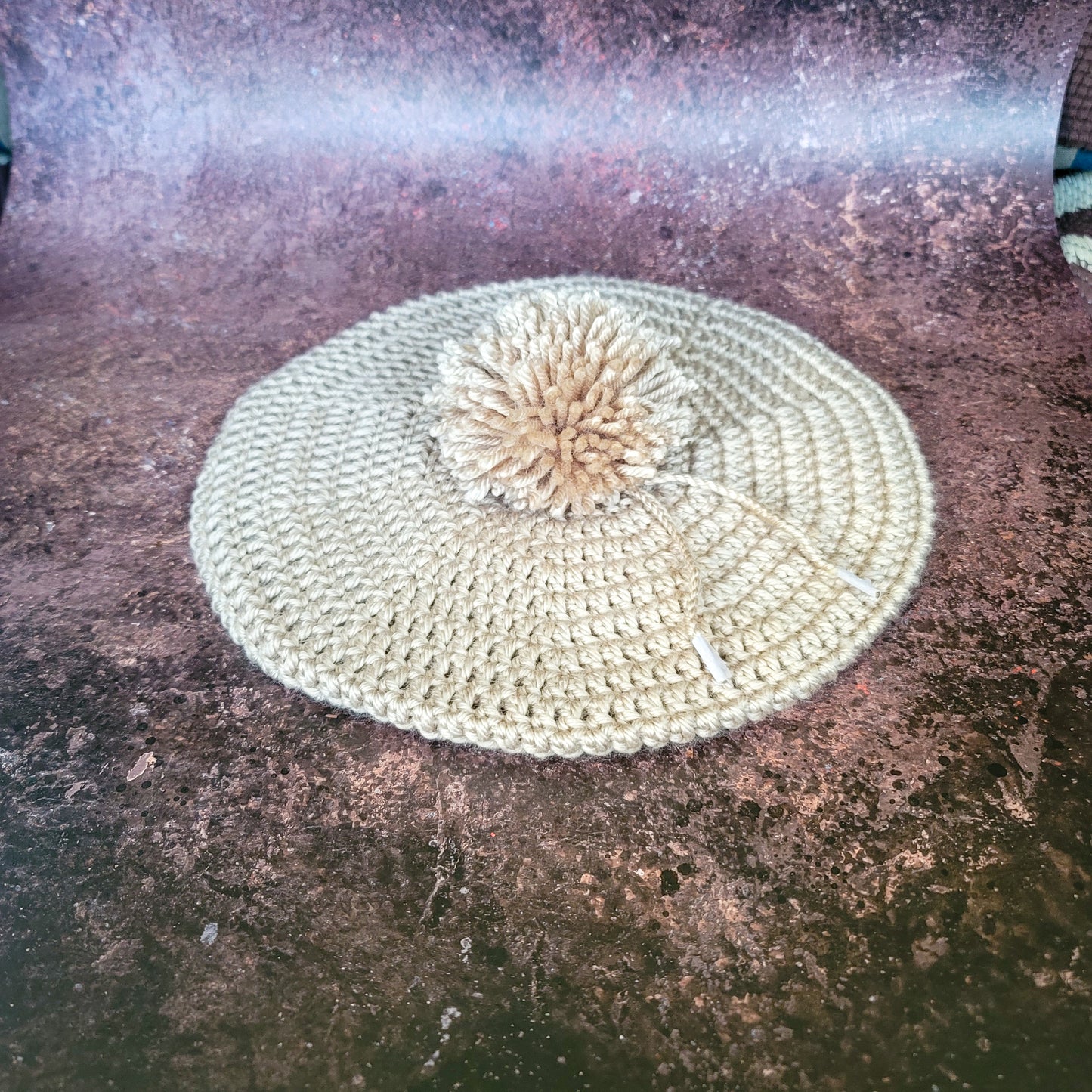 Tan crocheted beret with pom-pom on side, flat lay.